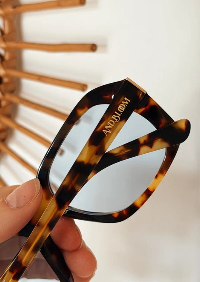 Phlox sunglasses product shot – hand holding tortoise frame with bold blue lenses, designed by AndBloom for women 35+